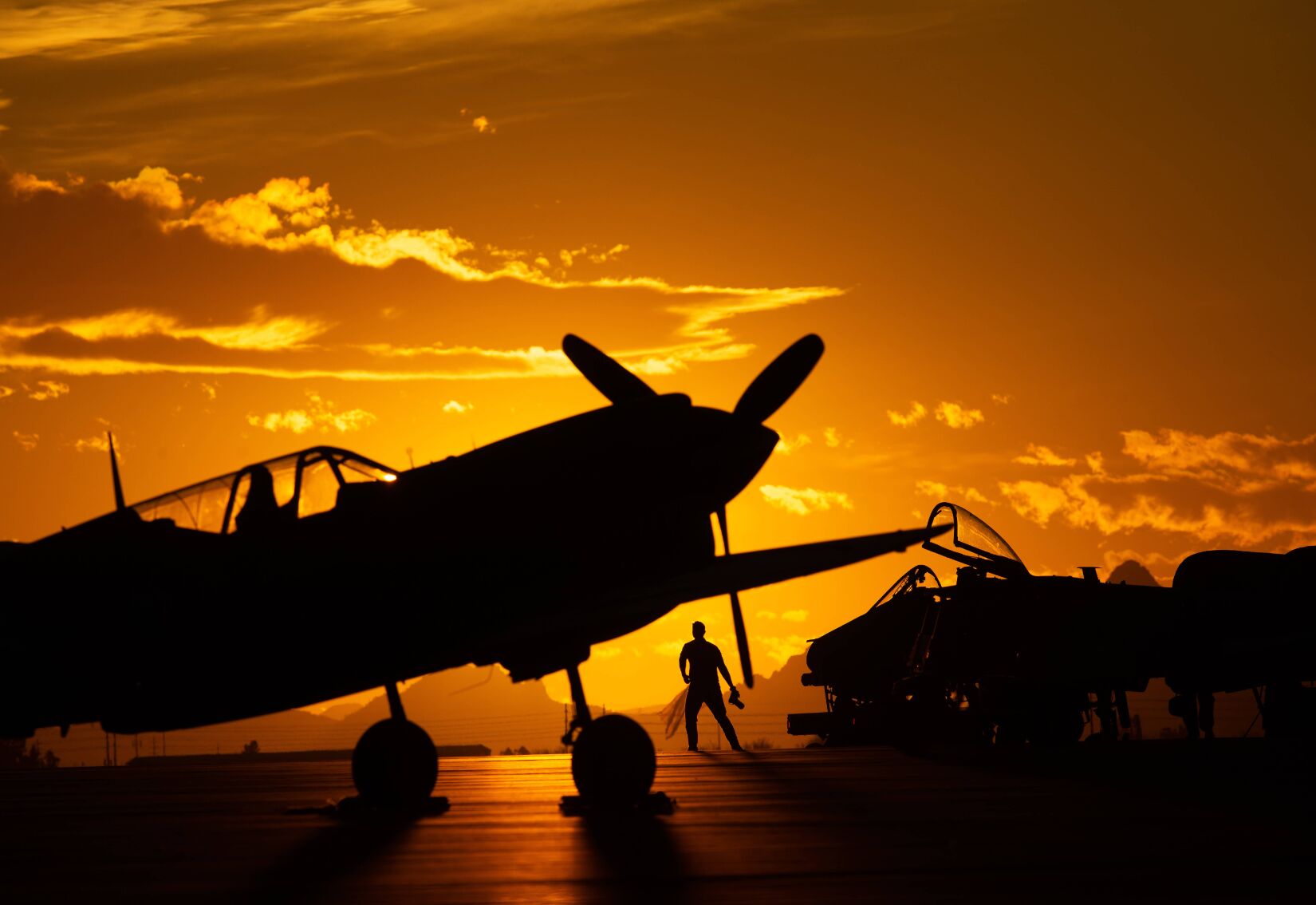 A propeller aircraft and an Air Force airman stand backlit against an orange sky.